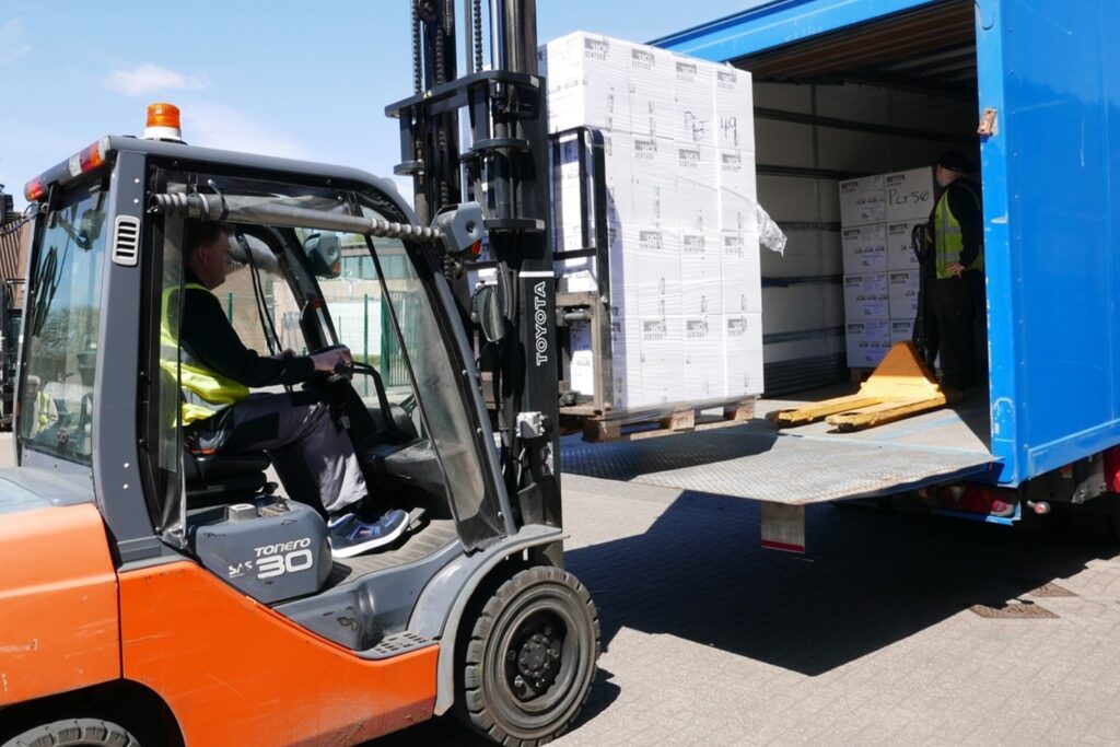 Two of FCDO Services' Logistics team loading supplies onto a blue lorry, one of whom is driving a fork lift truck. It is a sunny day