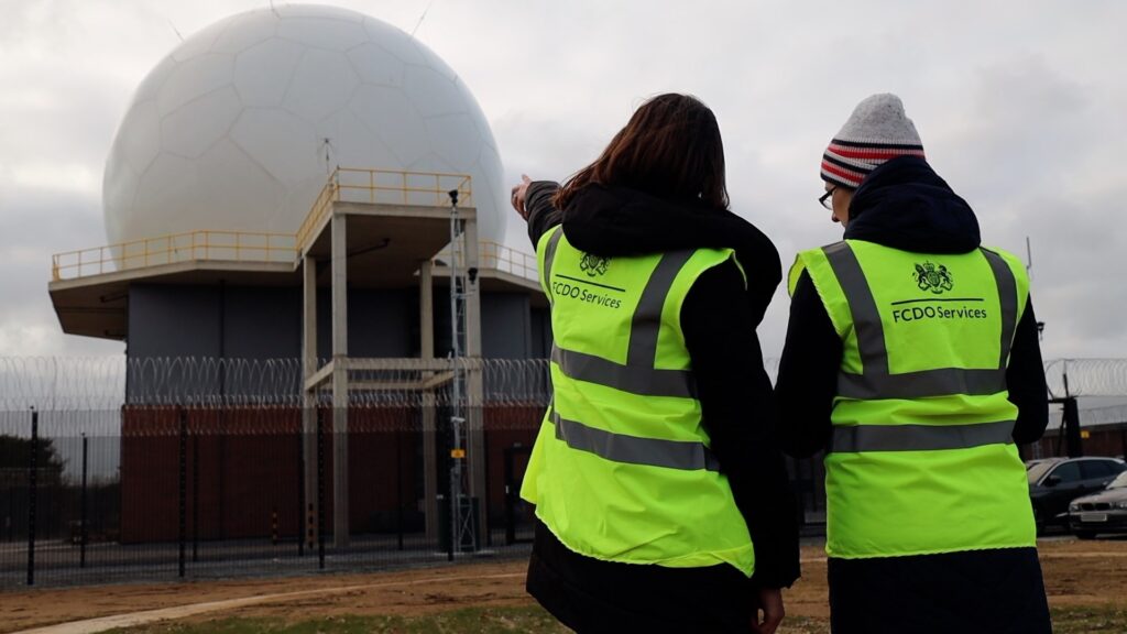 Two women in FCDO Services high vis vests, on a construction site pointing at an RAF radome.
