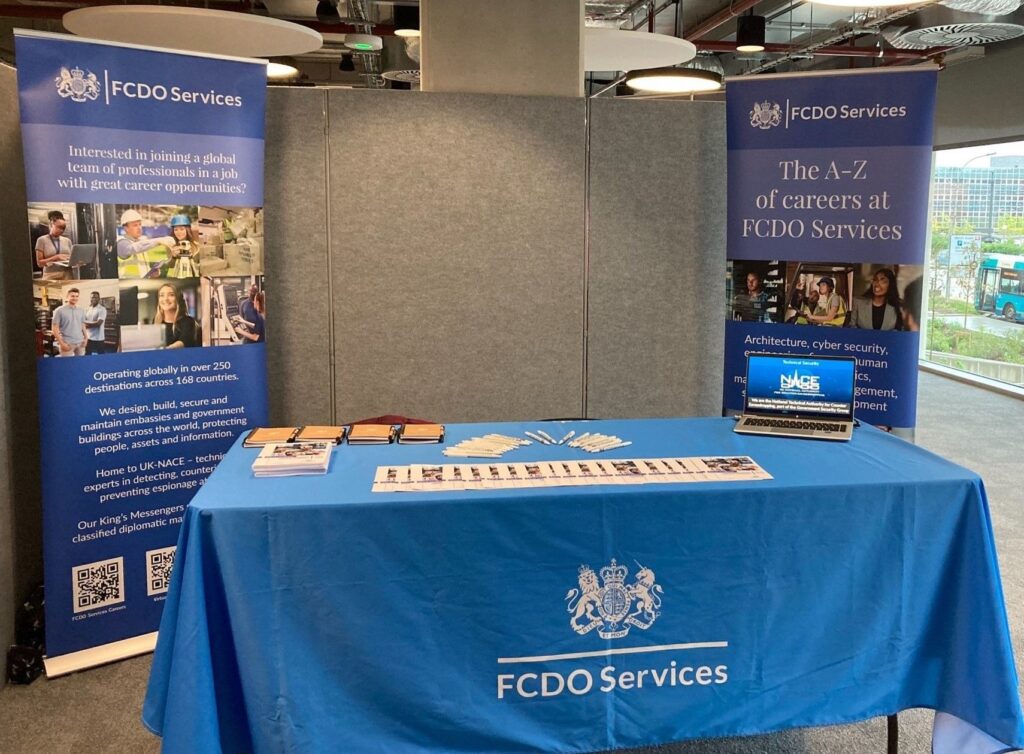Table with FCDO Services branded cover on it holding a laptop, leaflets and pens, in front of 2 pull up banners